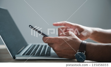 Close-up of a man's hands using a smartphone with a laptop in front of him. He's wearing a watch on his wrist. 135856033