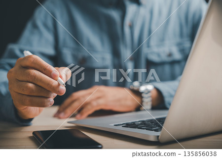 Close-up of a hand holding a stylus pen, with a laptop and smartphone in the foreground.  The man is wearing a blue shirt.  Perfect for business, technology, or productivity themes. 135856038