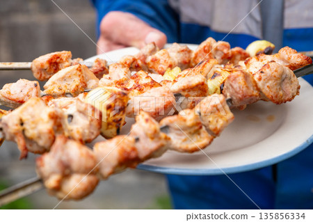 Man holding skewers of roasted pork shashlik over a plate. Traditional eastern european grilled meat dish for picnic, outdoor cooking food. Man holding skewers of roasted pork shashlik over a plate. Traditional eastern european grilled meat dish for picnic, outdoor cooking food. 135856334