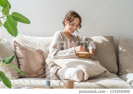 Smiling Asian woman sitting on the sofa in the living room and eating popcorn Smiling Asian woman sitting on the sofa in the living room and eating popcorn 135856900