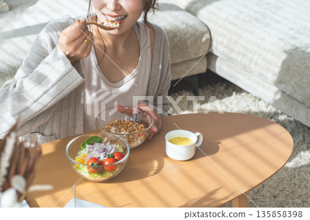一位女士坐在客廳的桌旁，吃著格蘭諾拉麥片和沙拉。 135858398