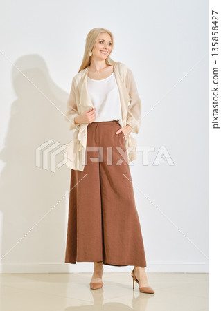 A smiling woman stands in a bright studio dressed in a light top and wide-leg brown pants. A smiling woman stands in a bright studio dressed in a light top and wide-leg brown pants. 135858427