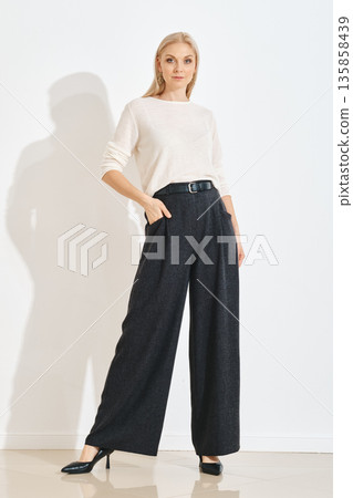 Woman stands confidently in stylish gray pants and white top in a simple indoor setting 135858439