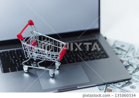Miniature Shopping Cart Rests On A Laptop Keyboard, With US Dollar Bills Scattered Nearby Miniature Shopping Cart Rests On A Laptop Keyboard, With US Dollar Bills Scattered Nearby 135860334