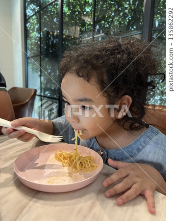 Cute little girl eating spaghetti with spoon in the kitchen in a restaurant. Healthy eating concept. Cute little girl eating spaghetti with spoon in the kitchen in a restaurant. Healthy eating concept. 135862292