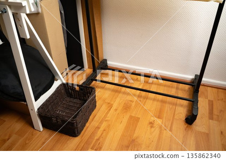Brown woven basket placed next to wooden trestles and a shoe rack in a casual home interior. Simple storage and organization. Concept of home, interior, lifestyle, domestic, minimal. Brown woven basket placed next to wooden trestles and a shoe rack in a casual home interior. Simple storage and organization. Concept of home, interior, lifestyle, domestic, minimal. 135862340