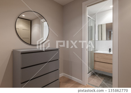 A modern bathroom interior featuring a gray dresser and a round mirror. The space is well-lit with a glass shower and wooden vanity. 135862770