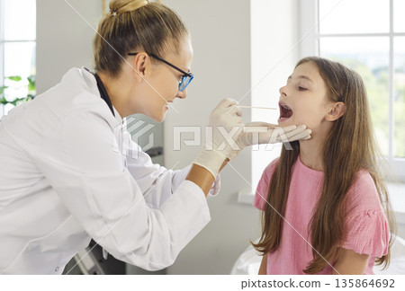 Pediatrician checking child throat with tongue depressor and swab in clinic 135864692