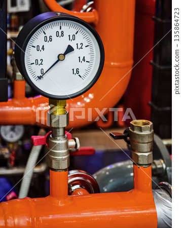 Pressure gauge on a bright orange pipe in an industrial setting with various equipment in the background during daytime Pressure gauge on a bright orange pipe in an industrial setting with various equipment in the background during daytime 135864723