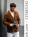 Stylish man in a brown suede blazer and white trousers leaning against a wall holding a small dark leather crossbody bag 135865342