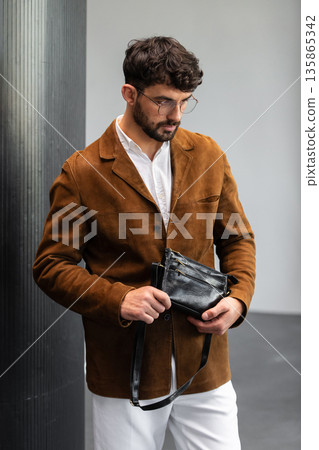 Stylish man in a brown suede blazer and white trousers leaning against a wall holding a small dark leather crossbody bag 135865342