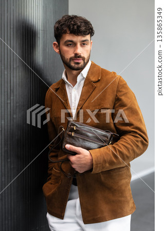 Stylish man in a brown suede blazer and white trousers leaning against a wall holding a small dark leather crossbody bag 135865349