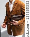 Stylish man in a brown suede blazer and white trousers leaning against a wall holding a small dark leather crossbody bag 135865352