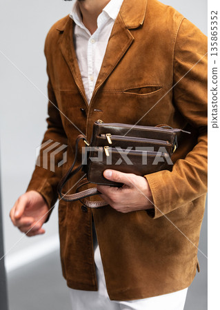 Stylish man in a brown suede blazer and white trousers leaning against a wall holding a small dark leather crossbody bag 135865352