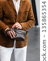 Stylish man in a brown suede blazer and white trousers leaning against a wall holding a small dark leather crossbody bag 135865354
