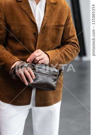 Stylish man in a brown suede blazer and white trousers leaning against a wall holding a small dark leather crossbody bag Stylish man in a brown suede blazer and white trousers leaning against a wall holding a small dark leather crossbody bag 135865354