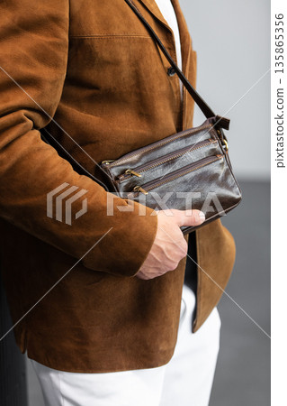 Stylish man in a brown suede blazer and white trousers leaning against a wall holding a small dark leather crossbody bag 135865356