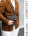Stylish man in a brown suede blazer and white trousers leaning against a wall holding a small dark leather crossbody bag 135865357