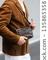 Stylish man in a brown suede blazer and white trousers leaning against a wall holding a small dark leather crossbody bag 135865358