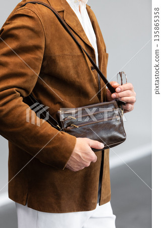 Stylish man in a brown suede blazer and white trousers leaning against a wall holding a small dark leather crossbody bag Stylish man in a brown suede blazer and white trousers leaning against a wall holding a small dark leather crossbody bag 135865358
