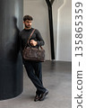 Confident man in sunglasses and charcoal knitwear holding a dark brown leather briefcase while leaning against a textured pillar in a modern minimalist interior. 135865359