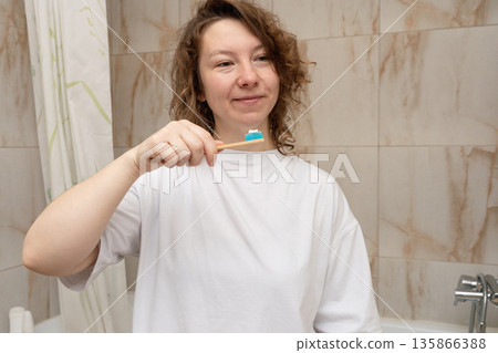 Woman holding a bamboo toothbrush with toothpaste, ready to begin her morning dental hygiene routine in the bathroom 135866388