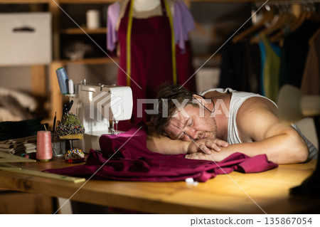 Late in evening, tired man tailor dozed off next to sewing machine. Late in evening, tired man tailor dozed off next to sewing machine. 135867054