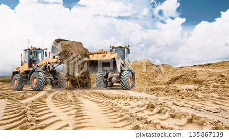 Two wheel loaders. Heavy equipment is working on sandy soil at a construction site. Track marks are visible in the sand, and a large pile of sand is visible in the background Two wheel loaders. Heavy equipment is working on sandy soil at a construction site. Track marks are visible in the sand, and a large pile of sand is visible in the background 135867139