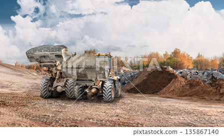 Loader works at construction site moving dirt and materials to a pile. The equipment operates on a busy ground with some trees in the background Loader works at construction site moving dirt and materials to a pile. The equipment operates on a busy ground with some trees in the background 135867140