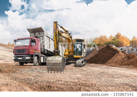 Loader moves earth while excavator digs at construction site. A truck is present, unloading materials near rental equipment under a clear sky and clouds Loader moves earth while excavator digs at construction site. A truck is present, unloading materials near rental equipment under a clear sky and clouds 135867141