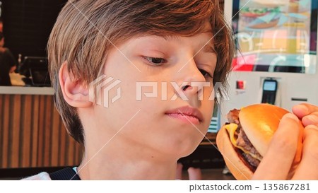 Cute boy eating burger. teenage boy eating a burger in a cafe. Hungry child biting burger in a restaurant. Fast food cafe. Cute boy eating burger. teenage boy eating a burger in a cafe. Hungry child biting burger in a restaurant. Fast food cafe. 135867281