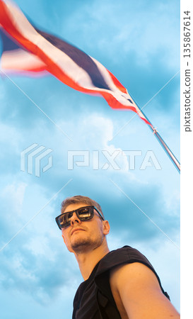 happy handsome tourist guy smiling with Thai flag at sunset, joyful man posing against Thailand flag evening sky, Asia travel and tourism 135867614