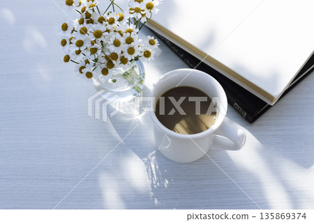 White table with sunlight filtering through the trees, coffee, matricaria, and a notebook (horizontal, blank space) White table with sunlight filtering through the trees, coffee, matricaria, and a notebook (horizontal, blank space) 135869374