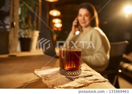 Coffee glass on table with woman talking on phone and laptop in background. Coffee glass on table with woman talking on phone and laptop in background. 135869463