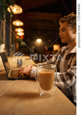 Double wall latte glass on table with man working on laptop. Double wall latte glass on table with man working on laptop. 135869466