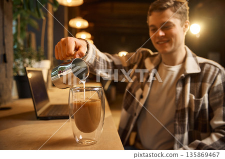 Man pouring milk into double wall glass coffee cup indoors. Man pouring milk into double wall glass coffee cup indoors. 135869467