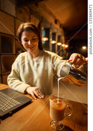 Woman smiling while pouring milk into coffee at wooden table. Woman smiling while pouring milk into coffee at wooden table. 135869471