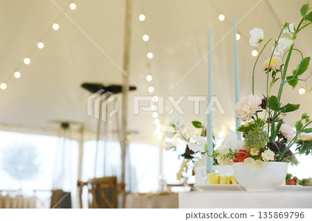 Elegant floral arrangement in a white bowl with pastel candles, set in a beautifully decorated tent for a special event, creating a romantic and inviting atmosphere 135869796
