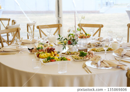 Table set with white tablecloth, plates of food, glasses, cutlery, floral centerpiece, and various dishes arranged on a round table in a tent during an event Table set with white tablecloth, plates of food, glasses, cutlery, floral centerpiece, and various dishes arranged on a round table in a tent during an event 135869806