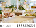 Platter of sliced meats and sushi rolls on white plates with greenery, crystal glassware, and floral decorations on dining table during event setup 135869834