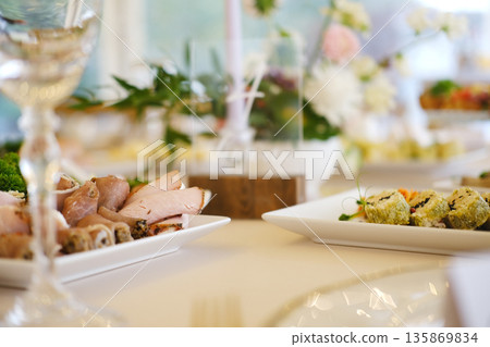 Platter of sliced meats and sushi rolls on white plates with greenery, crystal glassware, and floral decorations on dining table during event setup Platter of sliced meats and sushi rolls on white plates with greenery, crystal glassware, and floral decorations on dining table during event setup 135869834