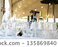 Glassware and wine bottles arranged on table under tent with string lights, featuring clear glasses, pitchers, and plates in outdoor setting during evening event 135869840