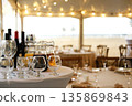 Table setting with wine bottles, glasses, and a pitcher on white tablecloth in a tent with string lights and wooden chairs during an outdoor event 135869843