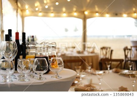 Table setting with wine bottles, glasses, and a pitcher on white tablecloth in a tent with string lights and wooden chairs during an outdoor event Table setting with wine bottles, glasses, and a pitcher on white tablecloth in a tent with string lights and wooden chairs during an outdoor event 135869843
