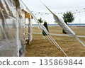 Tent with clear plastic walls and string lights set up on grass field with trees and pathway in background under cloudy sky during evening 135869844