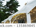 Tent securing process with ropes and stakes, ensuring stability and protection from wind, surrounded by lush trees and ambient lighting for outdoor events 135869845