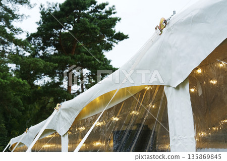 Tent securing process with ropes and stakes, ensuring stability and protection from wind, surrounded by lush trees and ambient lighting for outdoor events 135869845