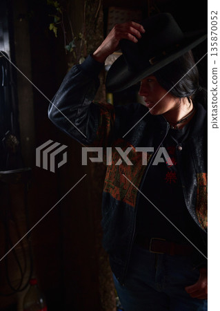 Cowgirl adjusting her wide-brim hat in a moody rustic setting with denim jacket 135870052