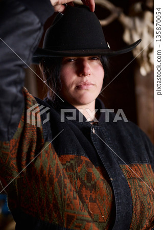 Cowgirl wearing a wide black hat and patterned jacket at a rustic ranch setting 135870054