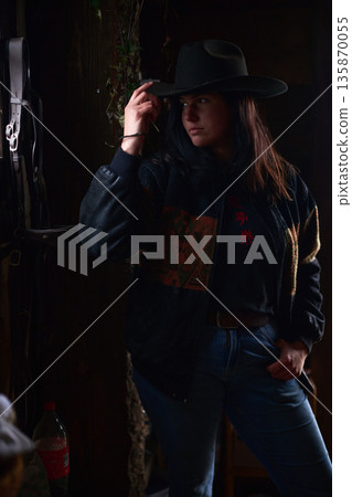 Cowgirl in a wide brim hat leans against a dark stable, exuding confidence at the ranch Cowgirl in a wide brim hat leans against a dark stable, exuding confidence at the ranch 135870055